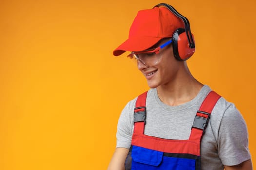 Young construction worker in helmet and uniform posing on yellow background in studio, close up