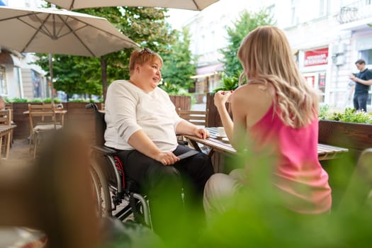 Mother with disability in wheelchair talking to her daughter while sitting at the table in cafe in the street