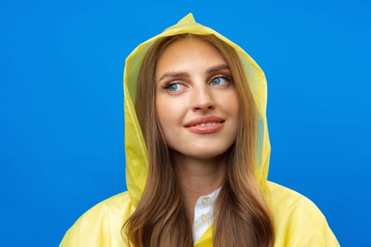 Young blonde woman wearing yellow raincoat smiling at camera over blue background in studio, close up