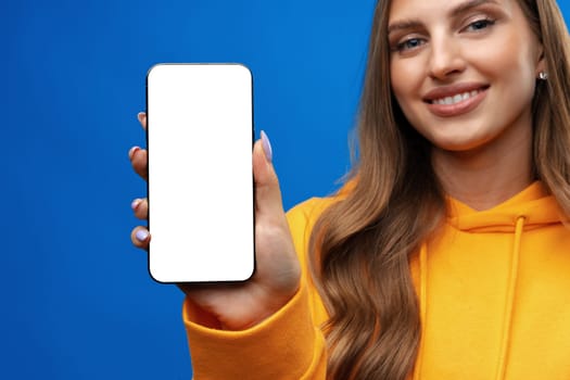 Young woman showing big blank cellphone screen on blue background in studio, copy space