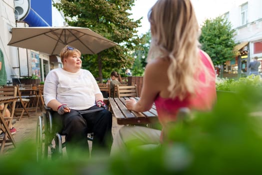 Mother with disability in wheelchair talking to her daughter while sitting at the table in cafe in the street