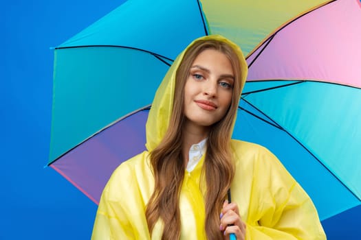 Young woman in yellow raincoat with rainbow umbrella against blue background in studio, close up