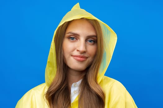 Young blonde woman wearing yellow raincoat smiling at camera over blue background in studio, close up