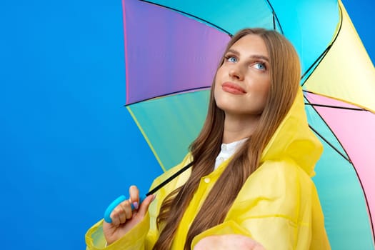Young woman in yellow raincoat with rainbow umbrella against blue background in studio, close up