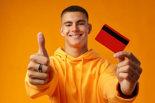 Handsome young man holding credit card on yellow color background in studio