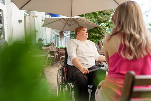 Mother with disability in wheelchair talking to her daughter while sitting at the table in cafe in the street