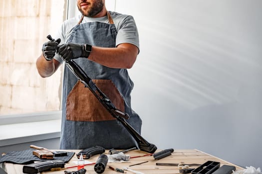 Close up of young man in apron disassembling a gun above the table in a workshop