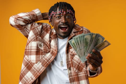 Young happy rich african man in casual shirt holding dollar bills against yellow background close up