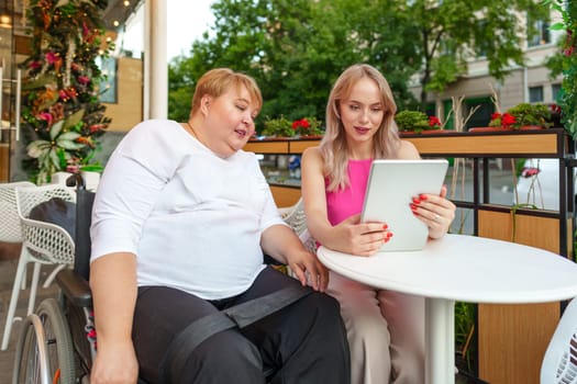 Mother with disability in wheelchair and her daughter using digital tablet while sitting at the table in cafe close up