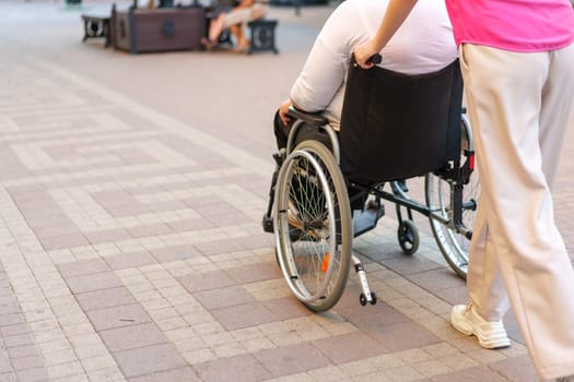 Back view of young woman helping mature woman in wheelchair strolling in the city