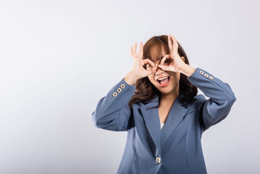 A cheerful young Asian woman stands gracefully, forming an 'OK' hand symbol near her eye, signifying agreement and positivity, in a studio with copy space.