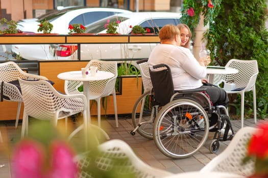 Young daughter and her mother in wheelchair sitting at the table in cafe with drinks and having fun, close up