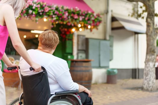 Back view of young woman helping mature woman in wheelchair strolling in the city