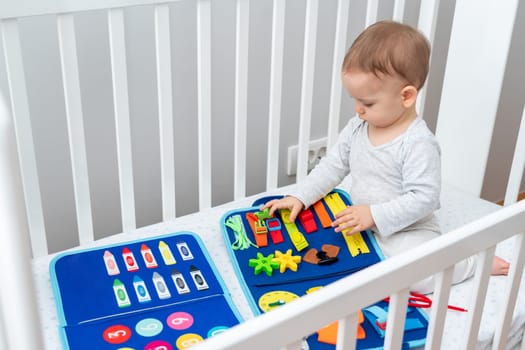 Little baby girl playing with busy book while sitting in crib. Concept of smart quiet books and modern educational toys