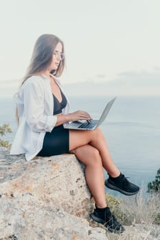 Digital nomad, woman in the hat, a business woman with a laptop sits on the rocks by the sea during sunset, makes a business transaction online from a distance. Freelance, remote work on vacation.