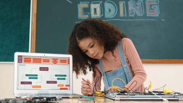 Smart girl inspect and fixing machine by using wire and connect system on table with laptop screen display engineering prompt or program generated AI while standing in front of blackboard . Pedagogy.
