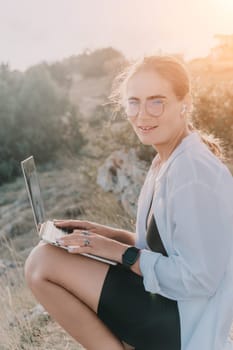 Digital nomad, woman in the hat, a business woman with a laptop sits on the rocks by the sea during sunset, makes a business transaction online from a distance. Freelance, remote work on vacation.