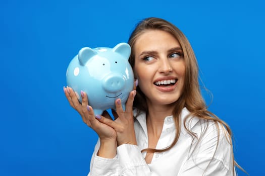 Young caucasian woman holding a piggy bank.against blue background in studio