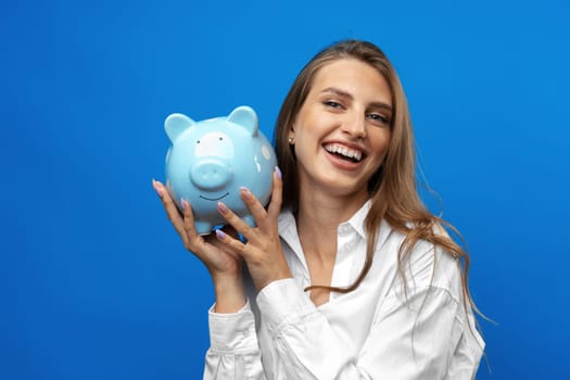 Young caucasian woman holding a piggy bank.against blue background in studio