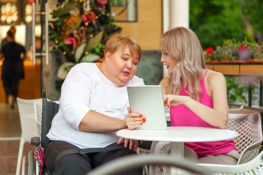 Mother with disability in wheelchair and her daughter using digital tablet while sitting at the table in cafe close up