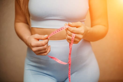 Cropped view of slim woman measuring waist with tape measure at home, close up. European woman checking the result of diet for weight loss or liposuction indoors.