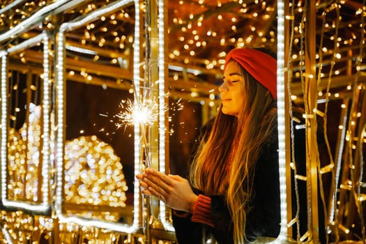 Woman holding sparkler night while celebrating Christmas outside. Dressed in a fur coat and a red headband. Blurred christmas decorations in the background. Selective focus.