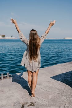 woman in a dress stands on a dock by the water, her arms raised in the air. Concept of freedom and joy, as the woman is celebrating or expressing her happiness