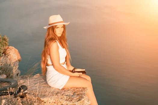 A tourist woman is sitting by the sea in a hat and white summer clothes, looking happy and relaxed