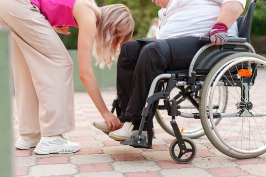 Young unrecognizable female helping woman patient with disability sitting on wheelchair in the street