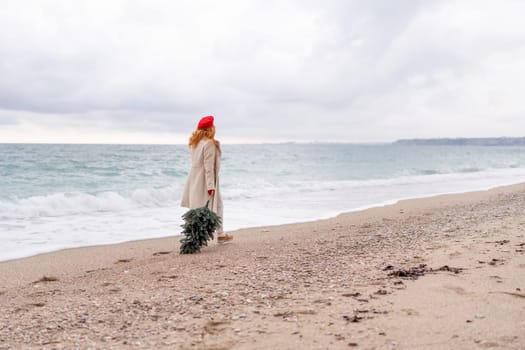 Redhead woman Christmas tree sea. Christmas portrait of a happy redhead woman walking along the beach and holding a Christmas tree in her hands. She is dressed in a light coat and a red beret