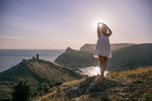 A blonde woman stands on a hill overlooking the ocean. She is wearing a white dress and she is enjoying the view