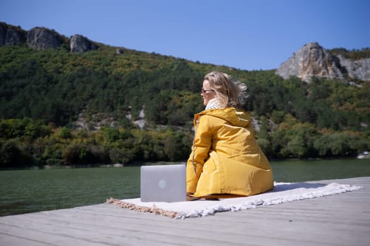 A woman in a yellow jacket sits on a rug by a lake with a laptop in front of her. Concept of relaxation and solitude, as the woman enjoys her time by the water while working on her laptop