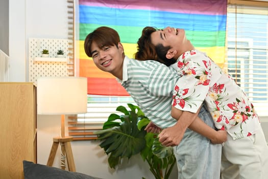 Laughing gay male couple giving piggyback ride for each other in living room with rainbow LGBT pride flag.