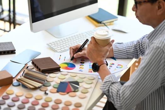 Senior male Interior designer drinking coffee and working on new design project at desk.