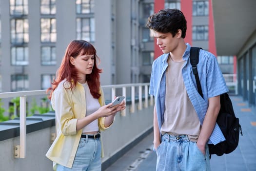 Friends young guy and girl university college students talking together, looking at smartphone outdoor, city modern buildings background. Lifestyle, communication, youth 19-20 years old, urban style