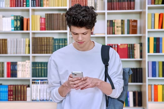 Teenage male student with backpack using smartphone, inside college building, in library. Technologies, mobile educational apps applications, services, e-learning concept