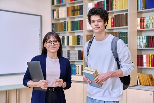 Female teacher standing with teenage college student guy looking at camera inside educational library office. Education, teaching, youth, lifestyle concept