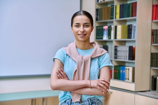 Portrait of smiling teenage girl student. Positive female teenager 16,17 years old with crossed arms inside high school building, background of library shelves books. Education, adolescence, lifestyle