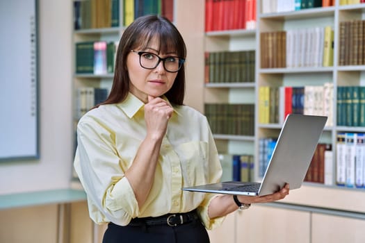 Middle aged elegant woman using laptop computer inside library. Confident serious elegant business female looking at camera teacher mentor professor leader manager psychologist counselor social worker