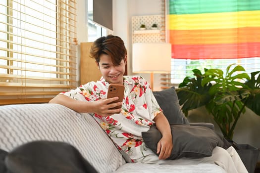 Smiling young man using mobile phone while sitting on couch in living room with rainbow LGBT pride flag.