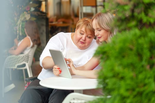 Mother with disability in wheelchair and her daughter using digital tablet while sitting at the table in cafe close up