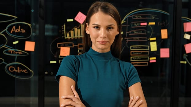 Professional businesswoman looking at camera while standing with arms crossing. Skilled beautiful worker crossing arm while standing in front of glass wall with mind map and sticky notes. Tracery