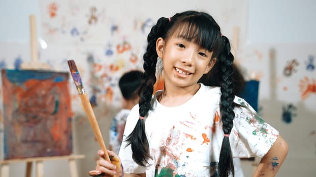 Creative cute girl pose at camera while diverse children painting behind. Young beautiful child looking at camera while standing at stained room and holding paintbrush. Creative activity. Erudition.