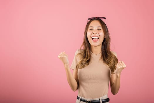 Happy Asian portrait beautiful cute young woman wear glasses makes raised hand up celebrating her winning success gesture, studio shot isolated pink background, Female excited say yes with copy space