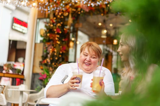 Young daughter and her mother in wheelchair sitting at the table in cafe with drinks and having fun, close up