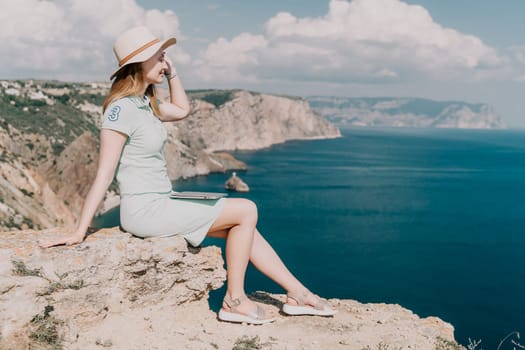 Digital nomad, woman in the hat, a business woman with a laptop sits on the rocks by the sea during sunset, makes a business transaction online from a distance. Freelance, remote work on vacation.