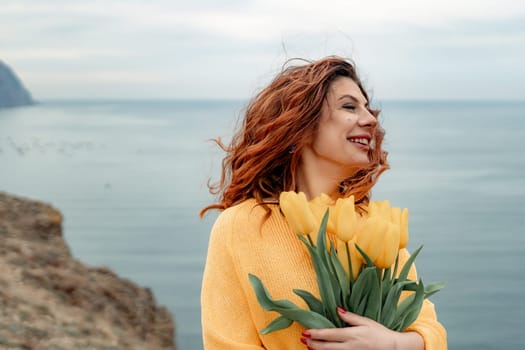 Portrait of a happy woman with hair flying in the wind against the backdrop of mountains and sea. Holding a bouquet of yellow tulips in her hands, wearing a yellow sweater.