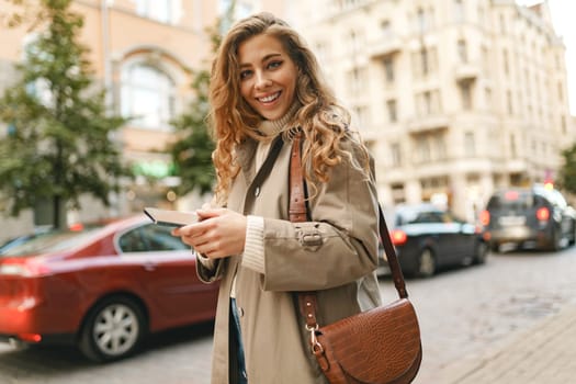 Smiling curly woman wearing warn coat walking down the street and using her phone, close up