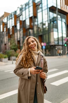 Smiling curly woman wearing warm coat walking down the street and using her phone, close up