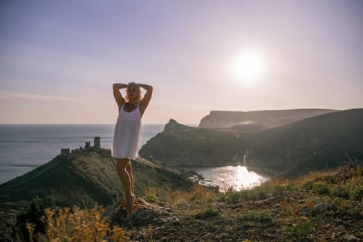 A blonde woman stands on a hill overlooking the ocean. She is wearing a white dress and she is enjoying the view